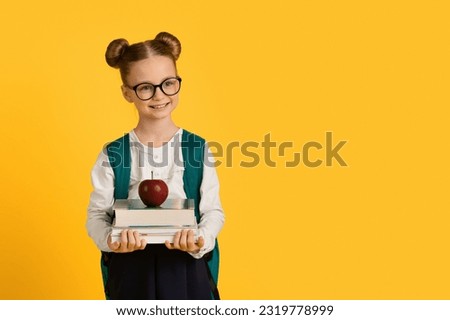 Education Concept. Little Cute Schoolgirl Holding Stack Of Books With Red Apple On Top, Smiling Nerdy Female Child With Backpack Posing Isolated Over Yellow Studio Background, Copy Space
