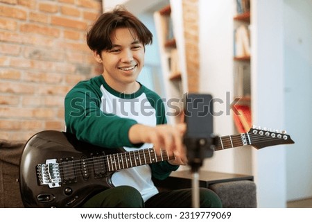 Talented Musician. Guy Playing Electric Guitar And Turning On Phone Camera Recording His Music For Online Audience At Home. Smiling Teenager Learning To Play Chords Via Internet. Selective Focus