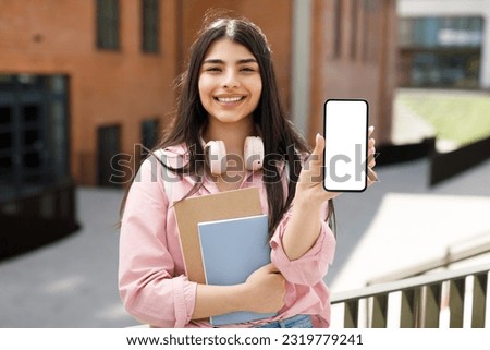 Happy teen student girl demonstrating blank smartphone and smiling, posing at college campus near university building outdoors, advertising educational app or website, mockup