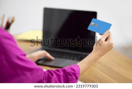 Online Shopping Concept. Over shoulder view of young woman holding credit card in hand and using laptop with black blank screen, unrecognizable female typing on keyboard, making purchases, mockup