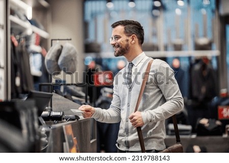 A cheerful young elegant man is using his credit card on a pos device in a store. Royalty-Free Stock Photo #2319788143