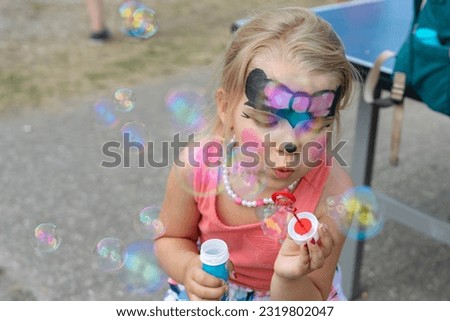 Little toddler girl having fun in a summer festival with face painting blowing bubbles . Royalty-Free Stock Photo #2319802047