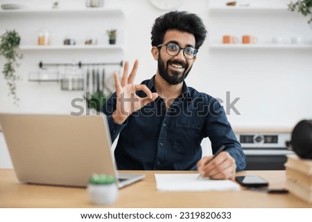 Portrait of attractive indian businessman posing at office desk with digital devices in spacious dining room, sign ok. Self-employed worker carrying out professional activity in comfortable workplace.