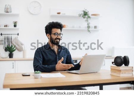 Young indian man in wireless headset conducting online meeting with teammates via laptop in kitchen interior. Serious freelance worker showing effective communication ability during web conference. Royalty-Free Stock Photo #2319820845