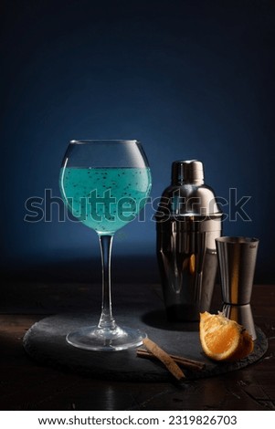 Bar composition on the table. Wine glass with a bright turquoise drink and an iron shaker. Orange slices and cinnamon sticks. Vertical frame. Royalty-Free Stock Photo #2319826703