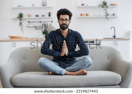 Young bearded man in jeans and dark shirt holding hands in anjali mudra while stretching in yoga pose on couch. Attractive indian adult relieving stress while sitting cross-legged in home interior. Royalty-Free Stock Photo #2319827443