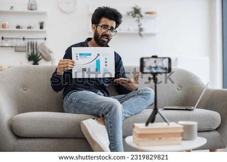 Enthusiastic lector of arab appearance in glasses teaching financial literacy via video link on cell phone. Intelligent professor showing currency growth graph to students during live streaming. Royalty-Free Stock Photo #2319829821