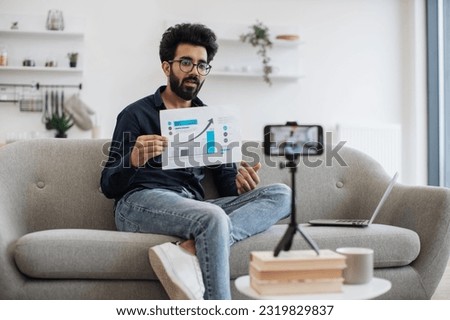 Enthusiastic lector of arab appearance in glasses teaching financial literacy via video link on cell phone. Intelligent professor showing currency growth graph to students during live streaming. Royalty-Free Stock Photo #2319829837