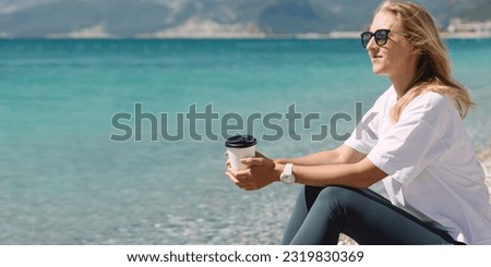 Pretty blond girl in white t-shirt holding coffee cup and enjoying beautiful view on seaside shore.  Blue mediterranean sea water on background with space for text