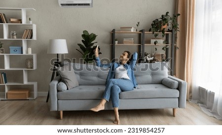 Happy young woman relax on couch in living room holding remote controller turn on air conditioner set comfort temperature, enjoy fresh air at summer day inside modern flat, ac, climate control concept Royalty-Free Stock Photo #2319854257