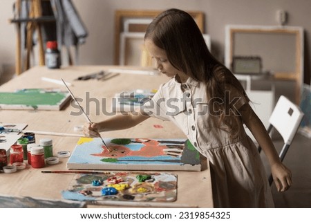 Cute pupil kid drawing cartoon animal in artist school classroom, using paintbrush, palette with acrylic paints, paper, canvas on big table, training creativity, art