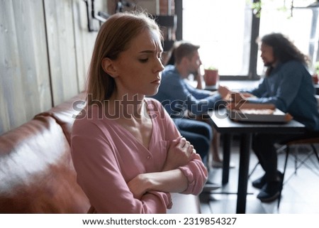 Unhappy abused young caucasian woman sitting on sofa in cafe separately from multiethnic diverse happy people, suffering from bullying, hurt by mockery gossip, unfair attitude or no friends support. Royalty-Free Stock Photo #2319854327