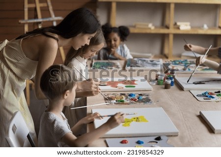 Multiethnic group of kids drawing cartoon animals on canvas, training creative talents on artistic class in school. Young teacher woman explaining blending acrylic colors on palette