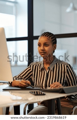 Young serious female designer with stylus and touchpad looking at computer screen and drawing graphic pictures while sitting by desk
