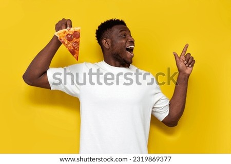 joyful african american man in white t-shirt holding piece of pizza and pointing finger to the side on yellow isolated background, the guy shows and advertises fast food Royalty-Free Stock Photo #2319869377