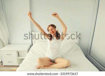 Happy cheerful young woman listening to music with wireless headphones having fun on bed in white room at home