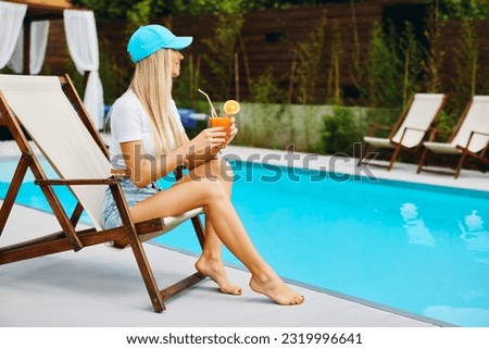 Attractive young woman sitting in a chair by the pool and drinking orange juice