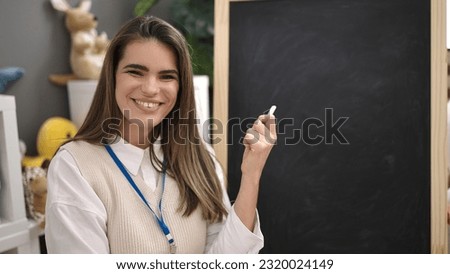 Young beautiful hispanic woman preschool teacher sitting by blackboard at kindergarten
