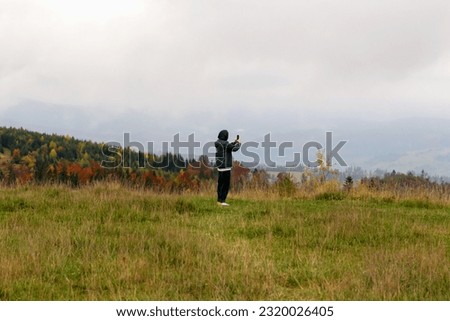 Unfamiliar man in cloudy mountains taking pictures with his phone. Beautiful autumn landscape, sky in clouds, grass and trees. Tourism and travel. Carpathian Mountains