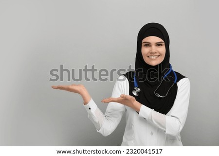 Muslim woman in hijab and medical uniform pointing at something on light gray background, space for text