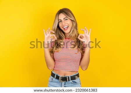 Young beautiful woman wearing striped t-shirt over yellow background  showing both hands with fingers in OK sign. Approval or recommending concept Royalty-Free Stock Photo #2320061383