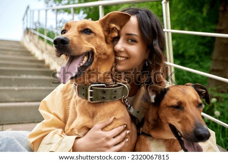 Young charming smiling girl is resting while walking in the park with two golden dogs. The girl hugs her pets. Love and affection between owner and pet. Adopting a pet from a shelter. Royalty-Free Stock Photo #2320067815