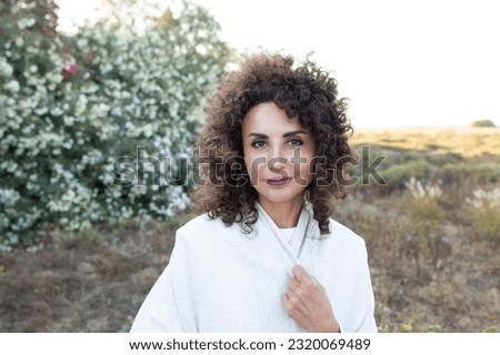 50 years old woman standing outside. Caucasian middle aged lady, curly brunette, relaxed. Wind blows the hair. Sunset light.Wellbeing, beauty, loneliness, people concept. Outdoor. Eye contact. Royalty-Free Stock Photo #2320069489