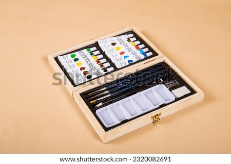 A landscape image of a water colour paint set on a beige background. The set is in a wooden box, there is a rainbow of colours, there are 3 brushes, a pencil, sharpener, eraser and a paint palette