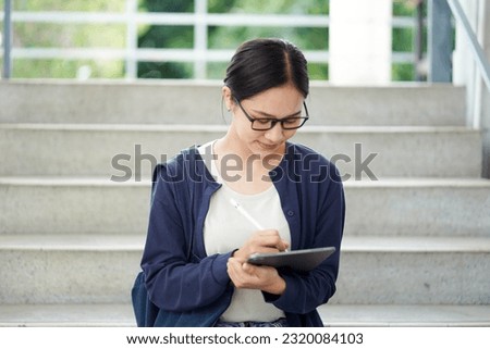 Closeup Asian young female student in casual cloth reading and make a short note on tablet for the exam on the stairs of the school building and blurred background. Asian school concept.