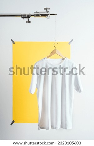Blank white t-shirt mockup, template on wooden hanger displayed on photo tripod over yellow paper backdrop sticked on the wall.