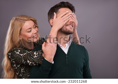 Half-length portrait of beautiful smiling blonde standing behind her handsome boyfriend closing his eyes. Isolated on dark background