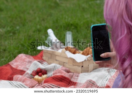 The girl takes pictures of a basket of champagne, croissants, glasses against the background of a plaid on green grass. Picnic in summer. Snack.