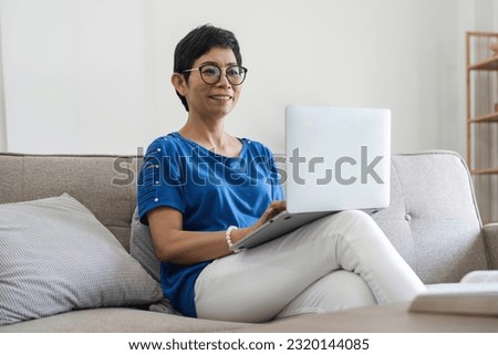 Happy successful mature business woman elderly working from home, holding laptop, looking at camera, smiling, resting in armchair, using modern technology, wireless connection for remote job