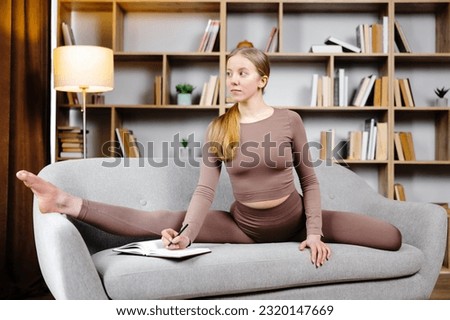 Young beautiful blonde woman in brown leggings and top sitting on a twine at home on the couch. The girl makes notes in a notebook while sitting on the sofa in an unusual position.