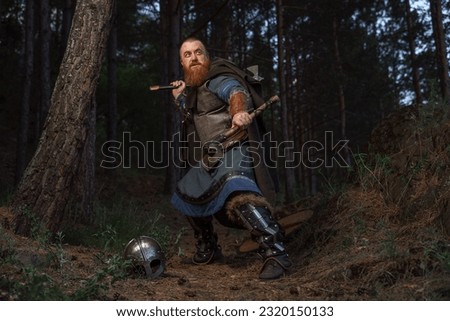 Medieval red-haired viking warrior with beard with two axes is preparing to attack on forest path.