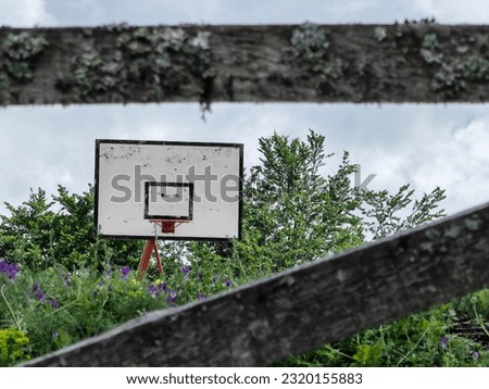 Framed white backboard, by wooden fence. Red hoop. Basketball rim. In the nature. White sky. No net.