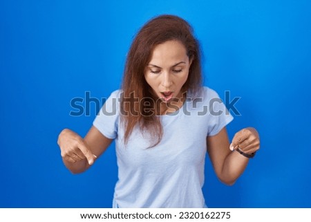 Brunette woman standing over blue background pointing down with fingers showing advertisement, surprised face and open mouth 