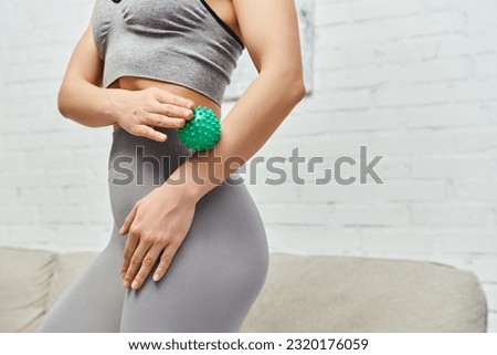 Cropped view of woman in sportswear massaging hand with manual massage ball while standing at home, balancing energy and holistic healing concept, myofascial release