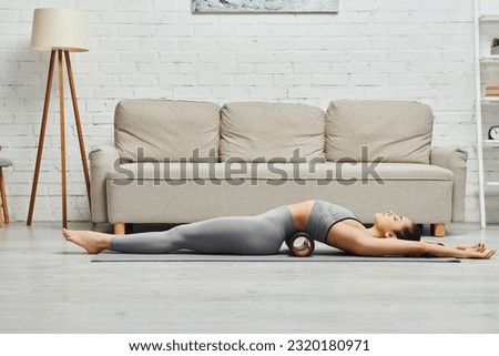 Side view of young brunette woman in activewear relaxing while lying on modern roller massager and fitness mat in living room at home, promoting lymph flow and wellness at home concept