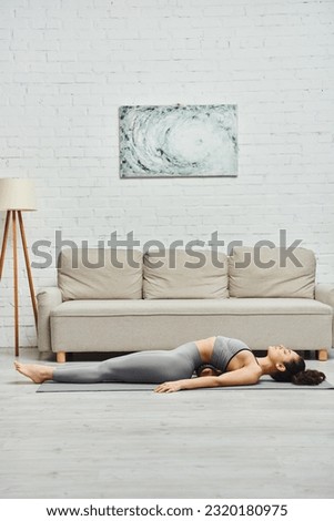 Side view of young brunette and barefoot woman in activewear lying on roller massager during self-massage on fitness mat at home, promoting lymph flow and wellness at home concept, tension relief