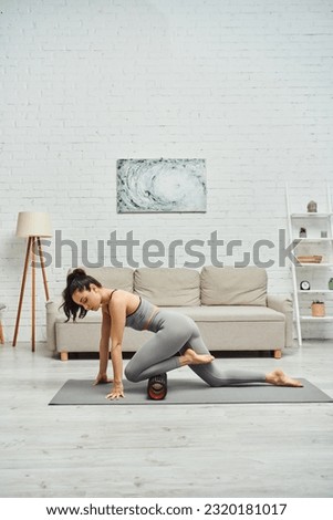Young brunette woman in activewear massaging knee with modern massage roller on fitness mat on floor in living room at home, sense of tranquility and promote relaxation concept, myofascial release