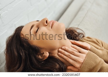 Close up view of relaxed brunette woman in brown jumper massaging neck during lymphatic drainage support and sitting on couch at home, self-care ritual and holistic healing concept, tension relief
