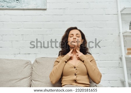 Young brunette woman with closed eyes in brown jumper touching neck during lymphatic nodes and circulation massage on couch at home, self-care ritual and holistic wellness practices concept