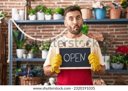 Handsome hispanic man working at florist holding open sign making fish face with mouth and squinting eyes, crazy and comical. 