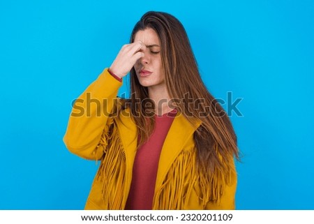 Very upset, young beautiful woman wearing jacket over blue studio background touching nose between closed eyes, wants to cry, having stressful relationship or having troubles with work