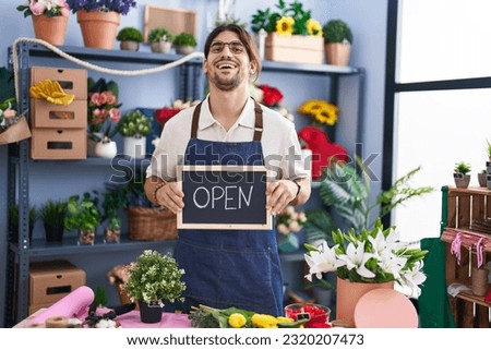 Hispanic man with long hair working at florist holding open sign smiling and laughing hard out loud because funny crazy joke. 