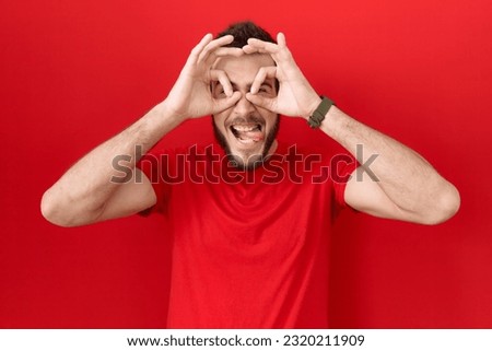 Young hispanic man wearing casual red t shirt doing ok gesture like binoculars sticking tongue out, eyes looking through fingers. crazy expression. 