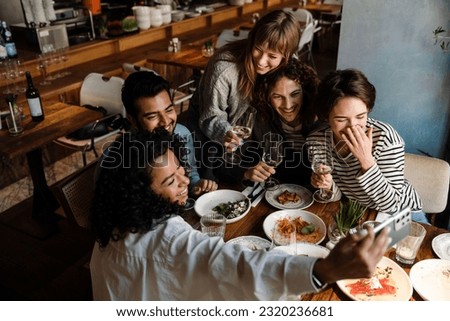 Group of young cheerful multinational friends taking selfie and drinking wine while dining at table in restaurant Royalty-Free Stock Photo #2320236681