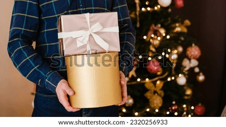 Men's hands holding gift boxes against the background of a Christmas tree. Gold and beige gift box for the holidays