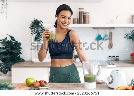 Shot of athletic smiling woman drinking smothie while listening music with earphones in the kitchen at home. Royalty-Free Stock Photo #2320266751